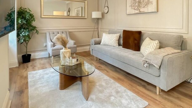 A living room with a grey sofa, a round glass coffee table, a potted plant, a framed mirror, and a wall clock. - Cozy 1 bed Blythswood Apartment (Glasgow City)