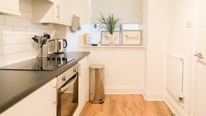 A compact kitchen with white cabinetry, a black countertop, a stainless steel kettle, and a small trash bin. - Cozy 1 bed Blythswood Apartment (Glasgow City)