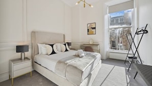 Un dormitorio con cama, mesitas de noche, una lámpara, radiador, ventana y un perchero.