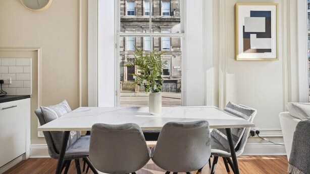 Un comedor moderno con una mesa blanca y dos sillas grises, una planta en maceta y vistas de un edificio a través de un ventanal.