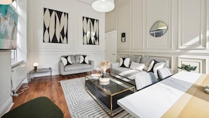 A modern living room with a sofa, a coffee table, and a dining table. There are abstract paintings on the wall and a mirror.