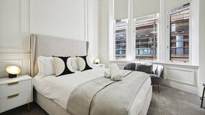 A neatly arranged bedroom with a large bed, bedside tables, a chair, and a window with a view of buildings.