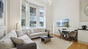 Un salón moderno con sofá, mesa de centro y sillones. Hay una mesa de comedor con sillas, un televisor montado en la pared y una ventana con andamios en el exterior.