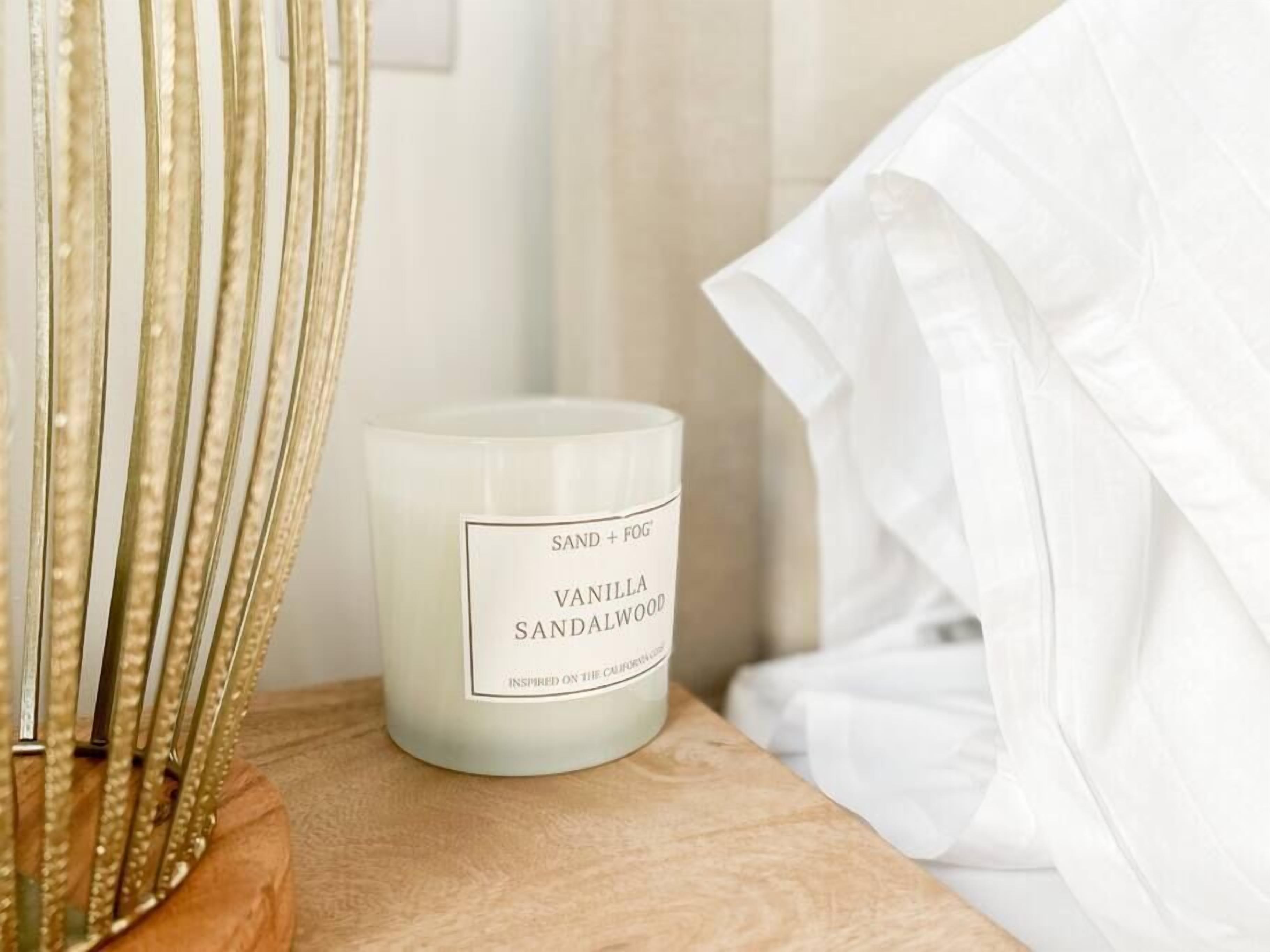 Una vela blanca con la etiqueta "SAND + FOG VANILLA SANDALWOOD" sobre una superficie de madera.