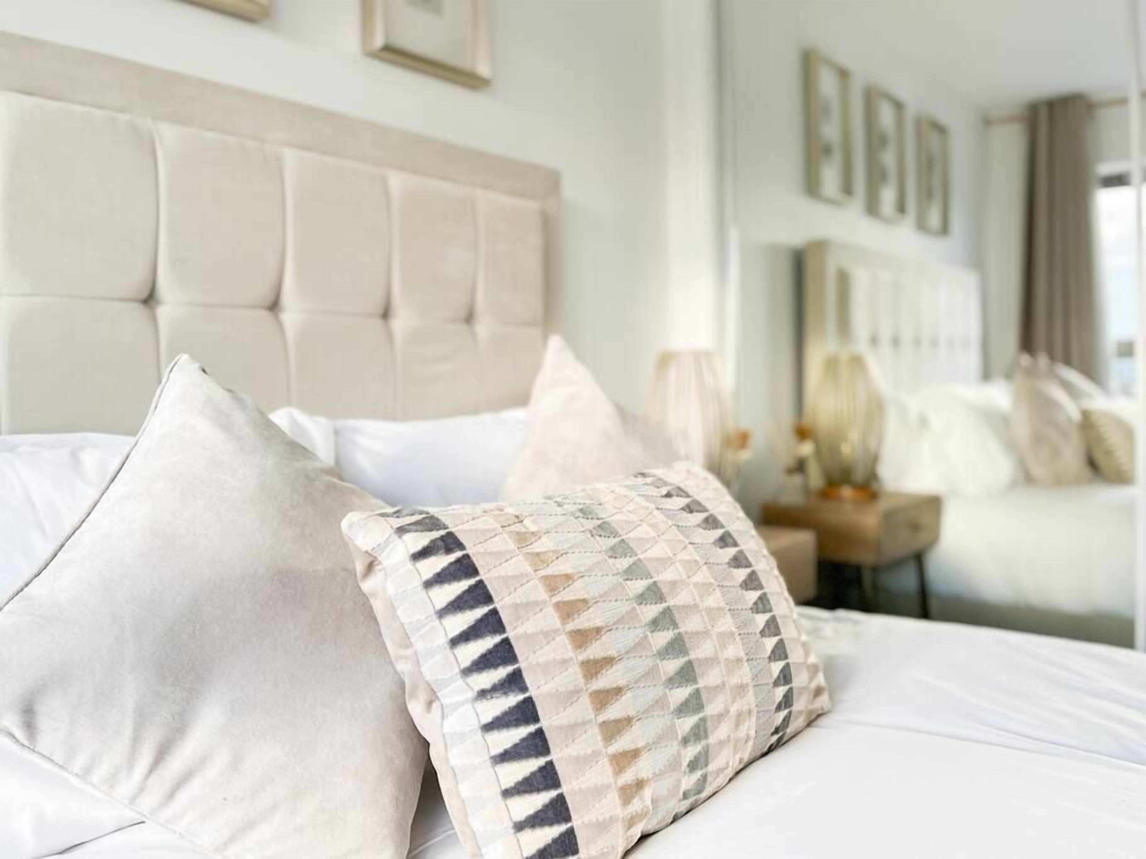 Un lit bien fait, avec du linge de lit blanc et deux coussins décoratifs, dont l’un présente un motif géométrique.