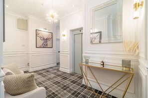 Um corredor com piso xadrez, uma poltrona branca, uma mesa de console dourada e uma obra de arte com um veado emoldurado.