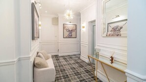 En gang med skakternet gulv, en hvid sofa med hynder, et billede af et dådyr i ramme, et glasmontre og et spejl med et billede i ramme.