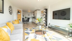 Een moderne woonkamer met een bank, salontafel en televisie. Achterin is een keuken met kasten en een eethoek.