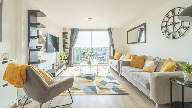 Een moderne woonkamer met een bank, fauteuil en salontafel, een televisie en een groot raam met gordijnen.