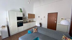 Een moderne keuken met witte kasten, een inbouwoven en een fornuis. Er is een grijze bank met blauwe kussens en een decoratief kussen. In de achtergrond is een houten deur en een radiator zichtbaar.