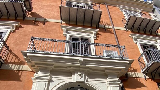 Een gebouw met een groot, versierd entreegebied, bestaande uit een dubbele deur met een metalen hek, een balkon met leuning en een bord dat de ingang aangeeft.