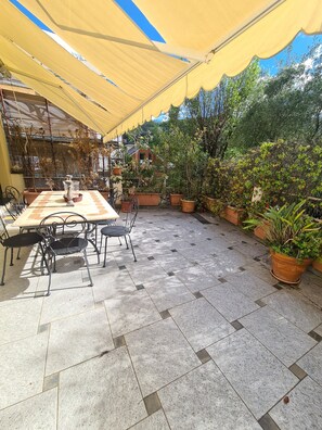 Pátio externo com mesa e cadeiras, rodeado por plantas em vasos e um toldo amarelo.