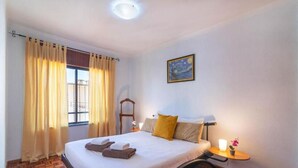 A hotel room with a bed, a bedside table with a lamp, a painting on the wall, and a window with curtains. - Quarteira Condestável T3 (Quarteira)