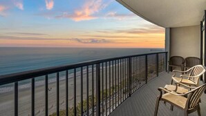 A balcony with a view of the ocean at sunset, featuring two wicker chairs and a railing.