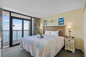 A hotel room with a large bed, a nightstand, a lamp, and a view of the ocean through a sliding door. - Compass Cove Resort Unit 1416 - Oceanfront (Myrtle Beach)