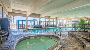 Une piscine intérieure avec un sol carrelé en pierre, un petit jacuzzi et un espace détente avec des chaises et des tables.