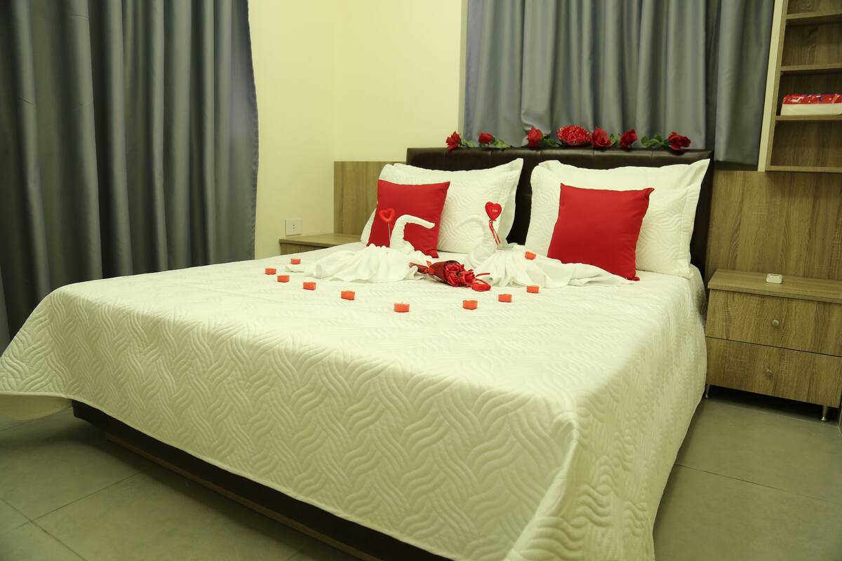 A neatly made bed with a white bedspread and red pillows, a wooden headboard, and a nightstand with a small table beside it.