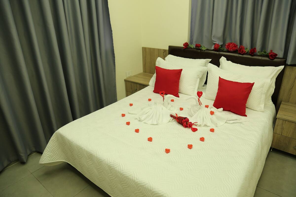 A neatly made bed with white bedding and red pillows, a wooden headboard with red roses, and a small wooden nightstand beside the bed.