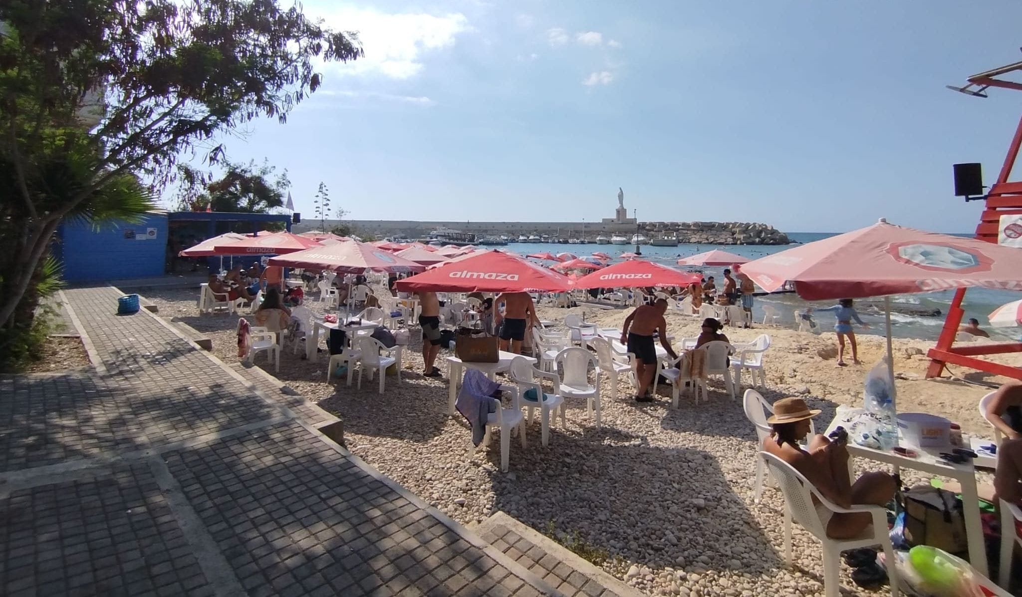 Un área de estar al aire libre con sombrillas rojas, sillas blancas y vista al mar y una estatua al fondo.