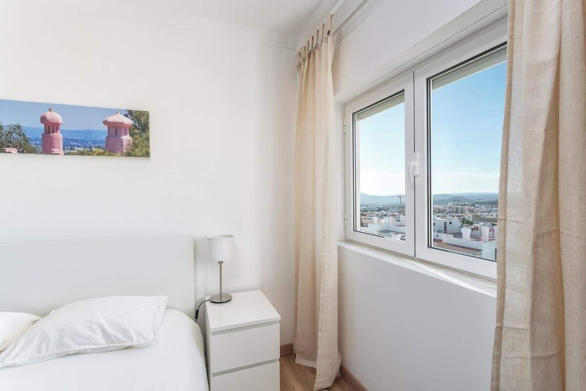 Un dormitorio con cama, mesita de noche con lámpara, ventana con cortinas y vista a la ciudad.