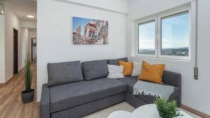 Un salon moderne avec un canapé d’angle gris, un tableau représentant un paysage urbain et une grande fenêtre offrant une vue dégagée.