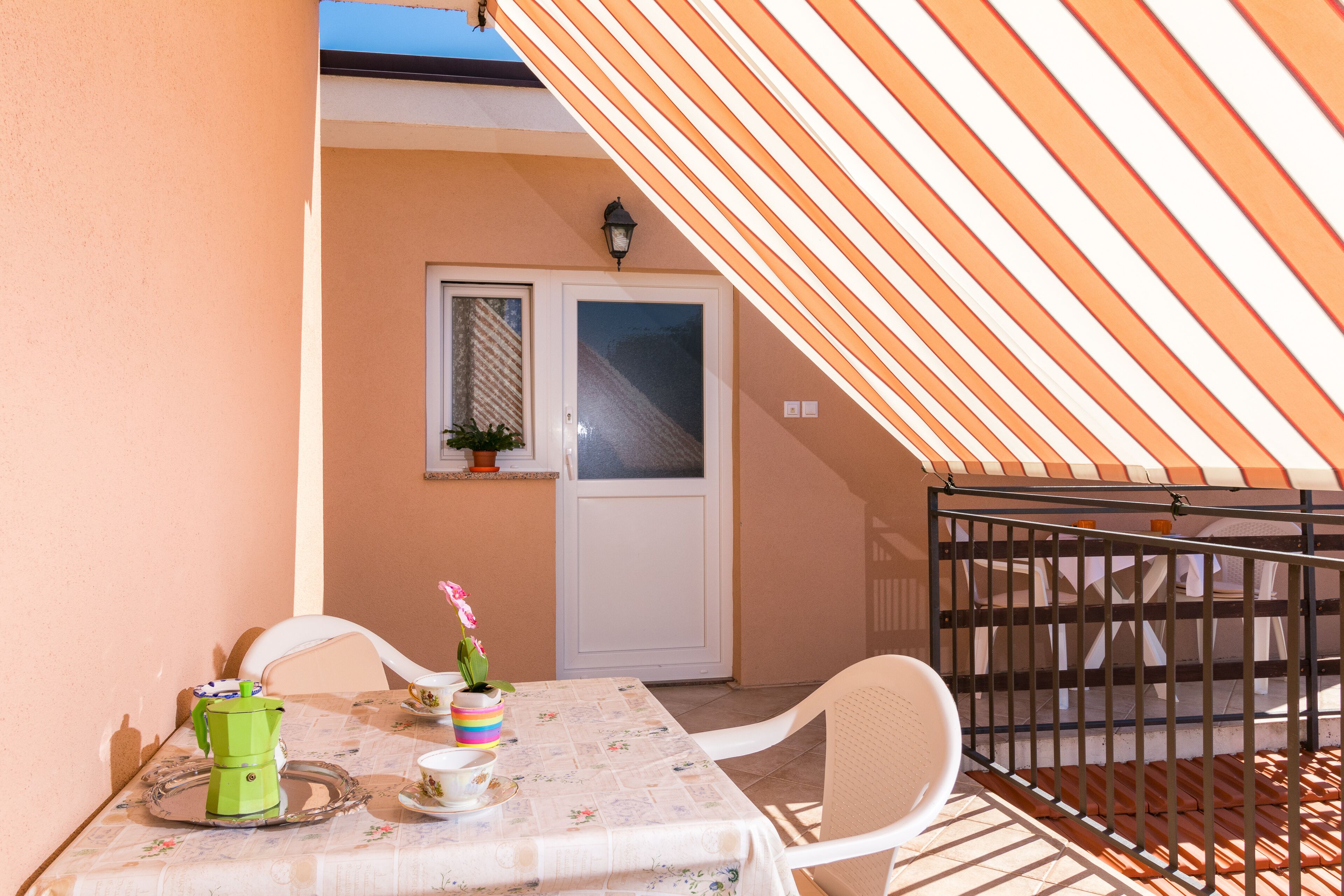 Un balcon avec un auvent rayé, une table recouverte d’une nappe à motifs floraux, une cafetière verte, une chaise blanche et une porte avec une fenêtre.