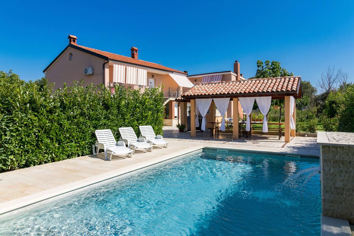 Ein Pool mit Liegestühlen, einer überdachten Sitzgruppe und einem Haus im Hintergrund.