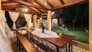 An outdoor patio with wooden tables and benches, a stone floor, and a view of a garden and house in the background. - Apartments Kendel (Fazana)