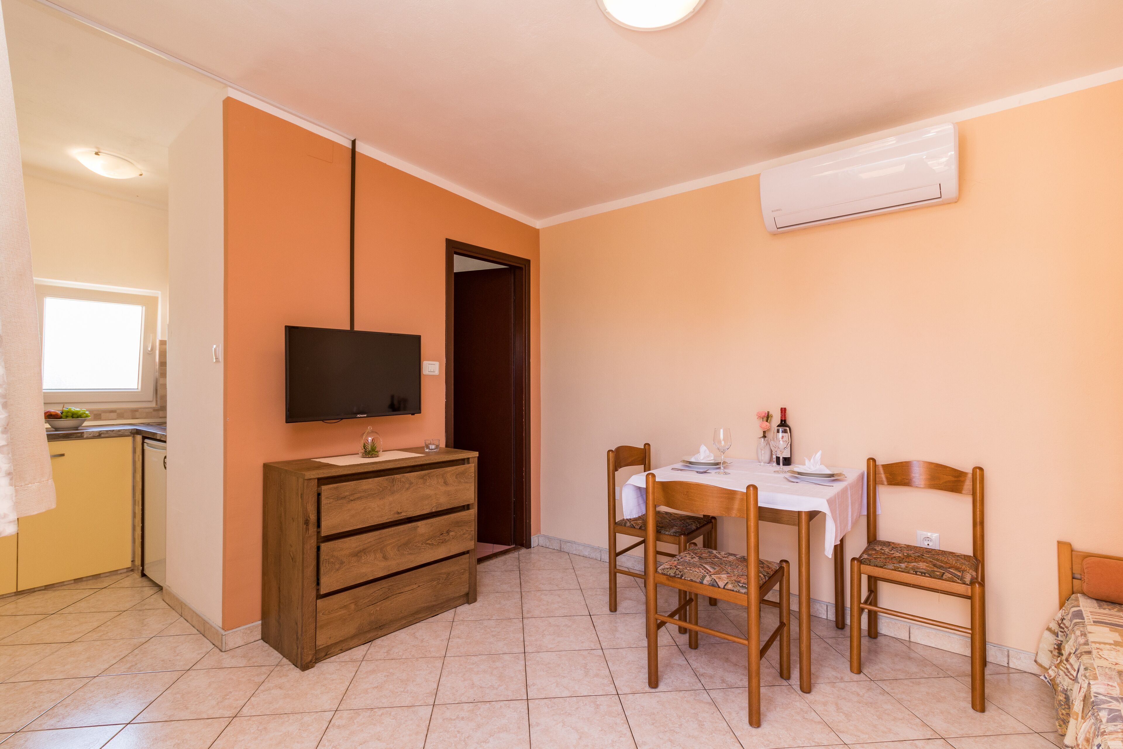 Un espacio habitable compacto con kitchenette, una zona de comedor con mesa y sillas, y un televisor montado en la pared.