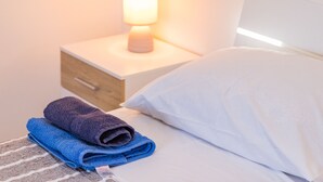 A neatly made bed with white and blue towels, a bedside lamp, and a wooden nightstand with a drawer. - Apartments Kendel (Fazana)