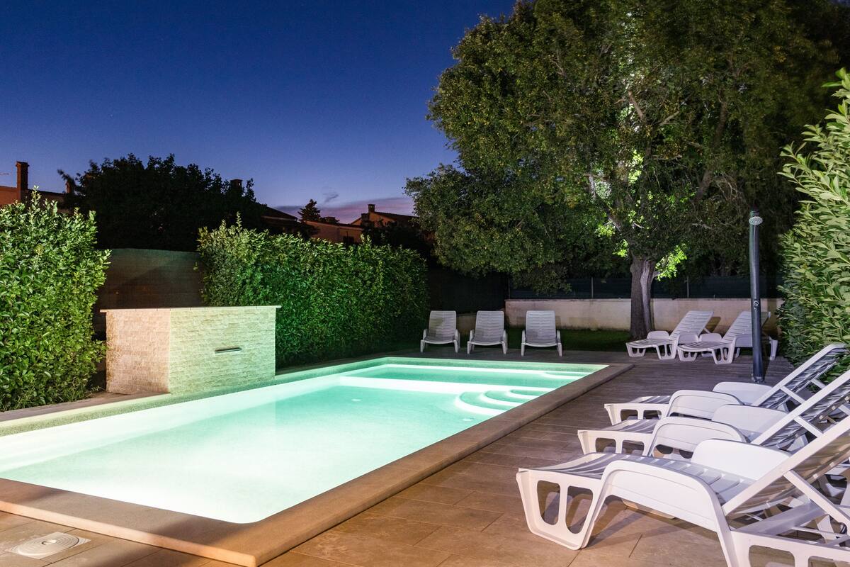 Ein rechteckiger Pool mit einer gefliesten Kante, umgeben von einer niedrigen Ziegelmauer und einem Hecken. Entlang des Poolrandes sind Liegestühle aufgestellt.