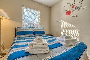 Quarto de hotel com cama, apresentando um edredom azul e branco com tema Nautica, um adesivo de parede do Mickey Mouse e toalhas dobradas com cuidado na cama.