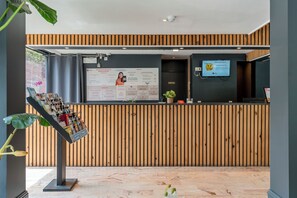 A reception desk with a wooden panel backdrop, a digital screen displaying information, and a plant on the counter. - Room-sleep & Go Brussels South - Budget (St. Genesius-Rode)