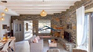 A cozy living room with a stone fireplace, a dining area with a table set for two, and a large window offering a view of the outside. - Mavrabeli Sunset Retreat, with amazing sea and sunset view, 15 minutes from port (Kéa)