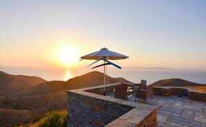 En terrasse med stenvægge og et bord med stole, med udsigt over et bjergrigt landskab i solnedgang.