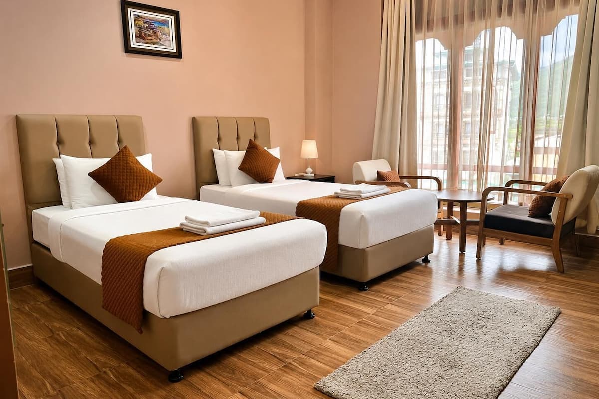 Habitación de hotel con dos camas, una mesita, una silla y vistas al exterior a través de amplias ventanas.
