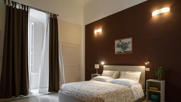 A bedroom with a bed, two bedside tables, a nightstand, a wall-mounted light fixture, a framed picture, and a door with curtains.