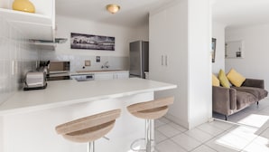 A modern kitchen with white cabinetry, a stainless steel refrigerator, and a white countertop. There are two woven stools at the kitchen island. - High Strand 38 by CTHA (Cape Town)