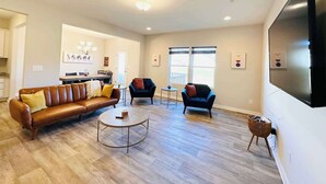 A spacious living room with a brown leather sofa, two armchairs, a coffee table, and a large flat-screen TV mounted on the wall.