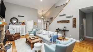 A spacious living room with a high ceiling, a staircase, and a cozy fireplace area. - House in the Woods: 4BR Harbert Retreat, 2 Fireplaces, Walk to Beach (Sawyer)