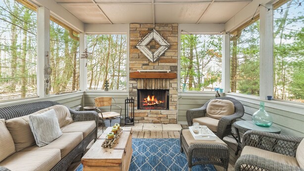 A cozy room with a stone fireplace, wicker furniture, and large windows overlooking a forest. - House in the Woods: 4BR Harbert Retreat, 2 Fireplaces, Walk to Beach (Sawyer)