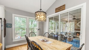 A dining area with a wooden table and chairs, a chandelier, and a view of an outdoor patio with a fire pit. - House in the Woods: 4BR Harbert Retreat, 2 Fireplaces, Walk to Beach (Sawyer)