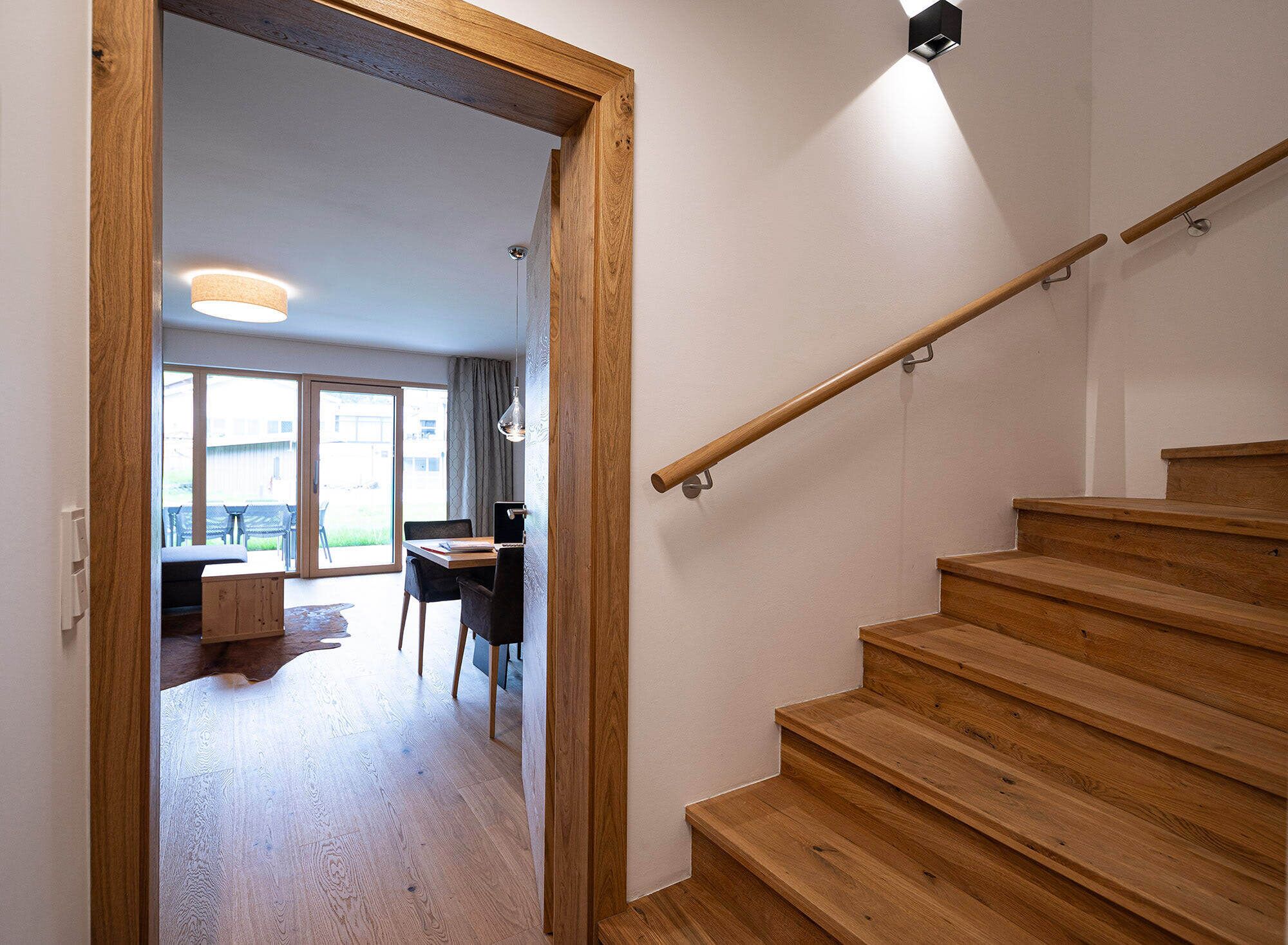 A wooden staircase leading to an upper floor, a dining area with a table and chairs, and a view of the outdoors through a glass door.