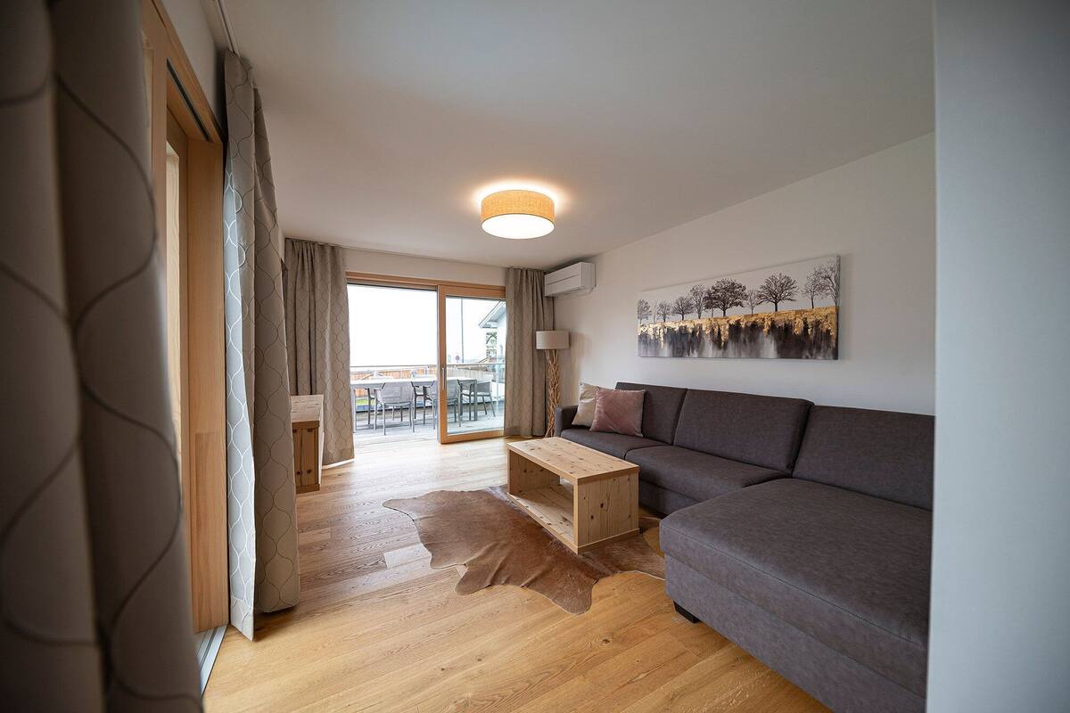 Ein modernes Wohnzimmer mit einem grauen Sofa, einem Holztisch und einem großen Fenster mit Blick auf einen Balkon.