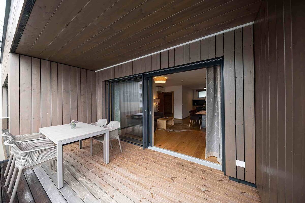 Eine Holzterrasse mit Tisch und Stühlen, eine Schiebetür zu einem Innenbereich mit Küche und Essbereich.