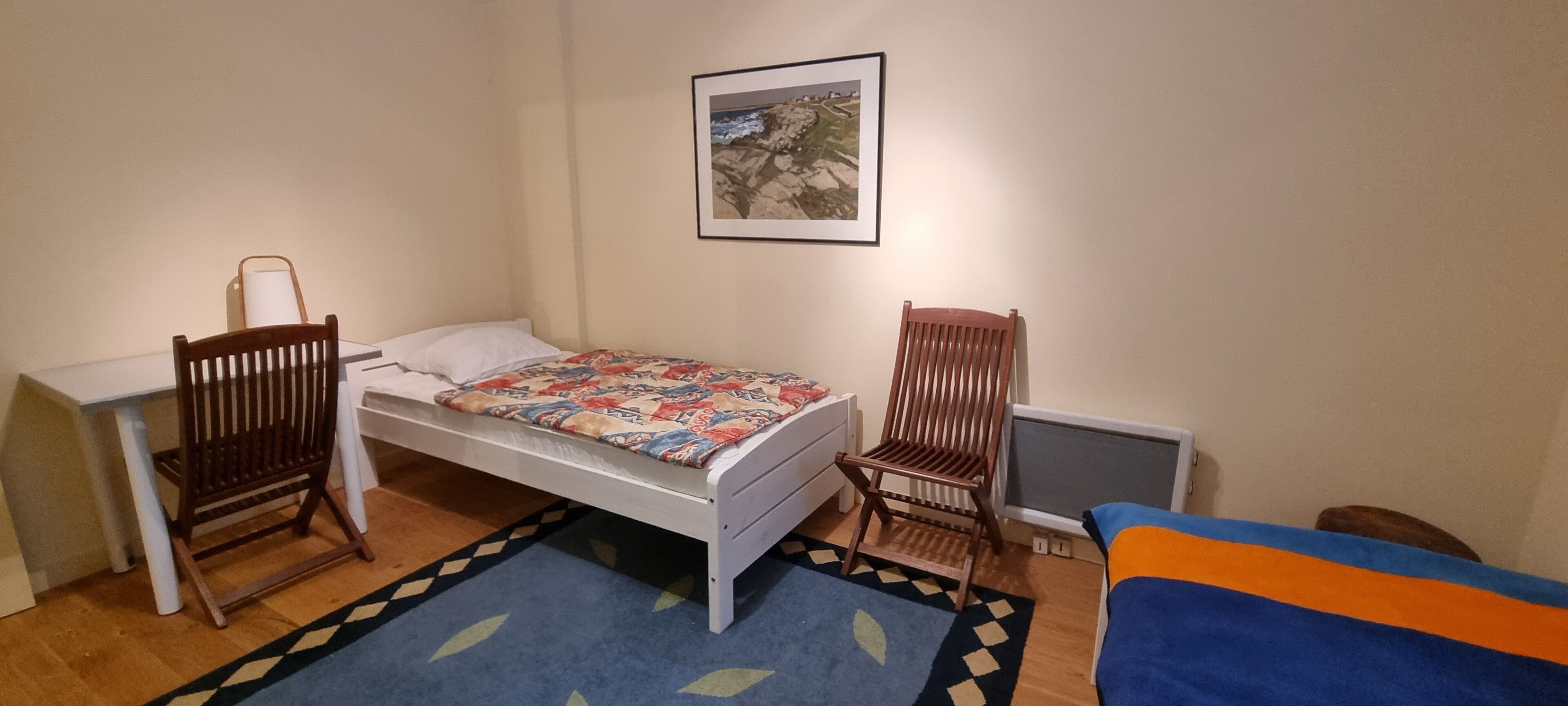 A small bedroom with a bed, a desk, two chairs, a radiator, and a framed picture on the wall.