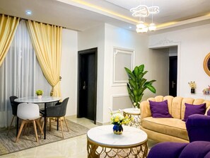 A modern living room with a round coffee table, a beige sofa with purple cushions, a dining table with chairs, and a large green plant. - Alexandria Court Lekki (Lekki)