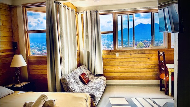 A hotel room with a bed, a sofa, a desk, and a chair. There are curtains on the windows, and a view of the outside.