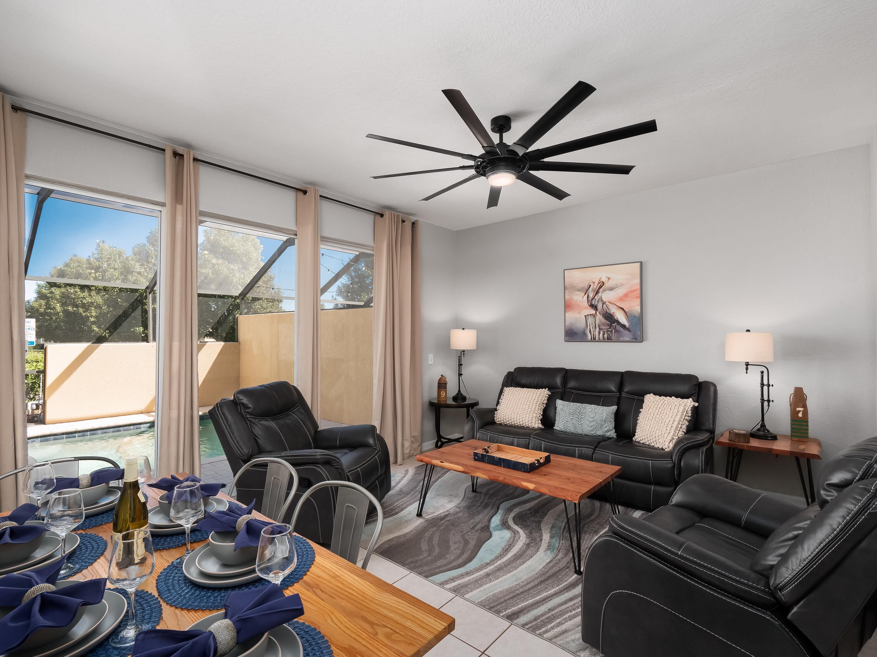 A modern living room with a black leather sofa, a wooden coffee table, and a dining area set with blue placemats and wine glasses.