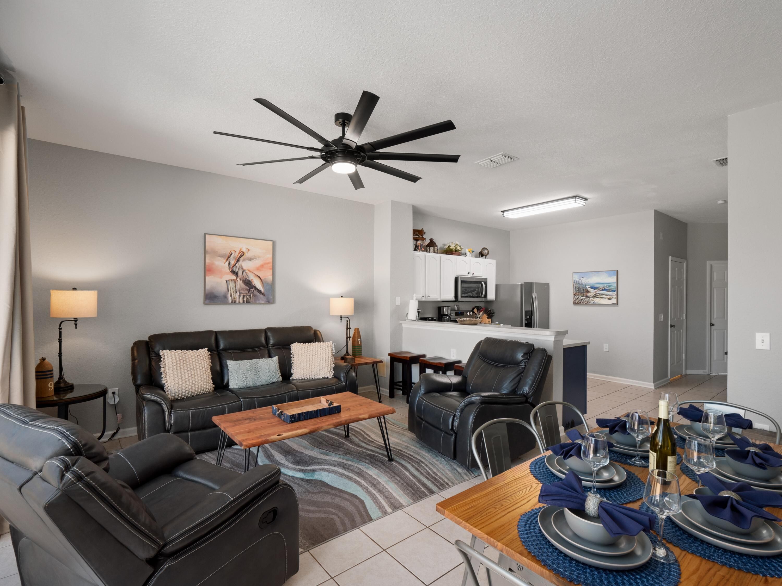 A modern living room with a black leather sofa, a wooden coffee table, and a dining area set for six with blue placemats and wine glasses.